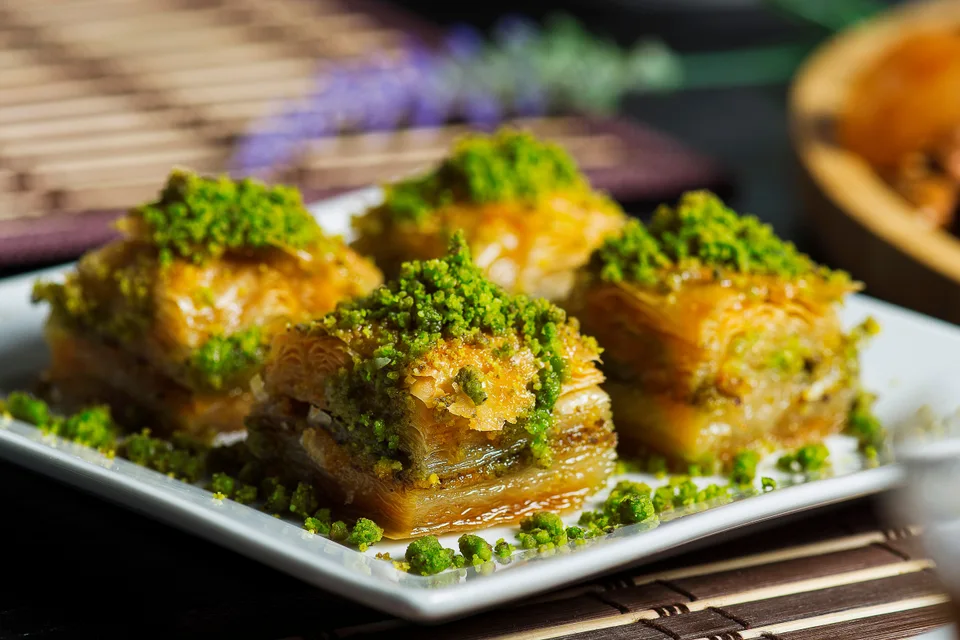 BookNoFees: Foto de ejemplo: Cuatro porciones de baklava en capas, cubiertas con pistachos triturados, en un plato blanco